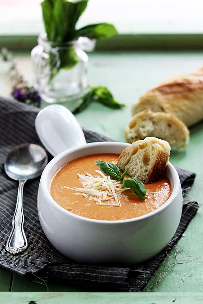 slow cooker tomato basil parmesan soup in a bowl with a piece of bread dipped in it with a spoon on the side and more bread and spices in the background.