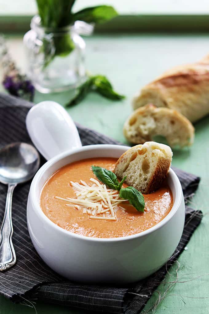 slow cooker tomato basil parmesan soup in a bowl with a piece of bread dipped in it and a spoon on the side with more bread and spices in the background.