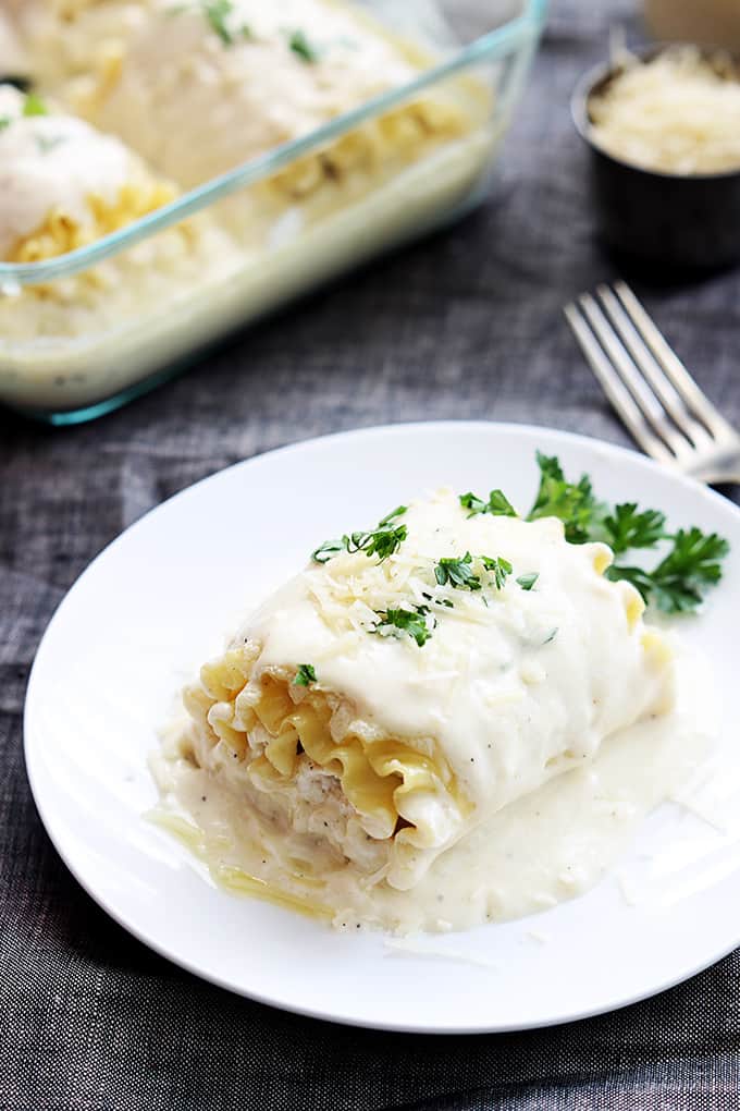 a white chicken lasagna rollup on a plate with a fork on the side and the rest of the rollups in a serving dish next to a small bowl of cheese faded in the background.