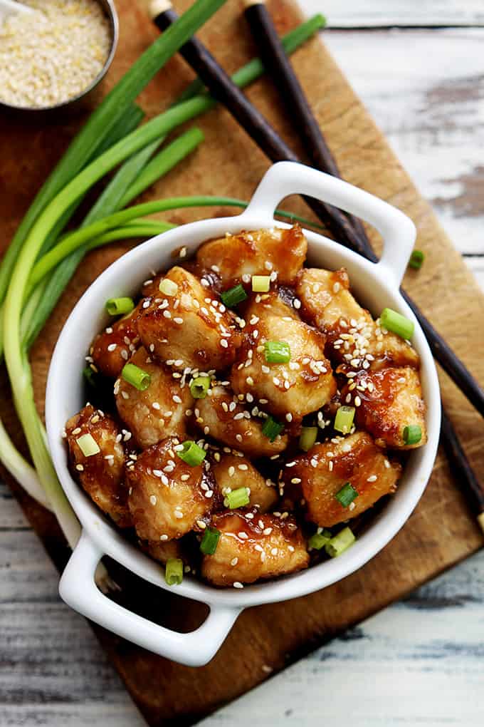 top view of baked sesame chicken in a bowl with chopsticks, green onions and a small bowl of sesame seeds on the side all on top of a wooden cutting board.