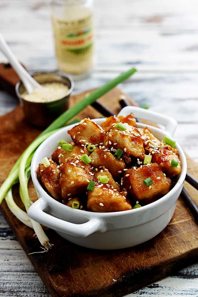 baked sesame chicken in a bowl with chopsticks and green onions on the side and sesame seeds in a bowl in the background all on a wooden cutting board.