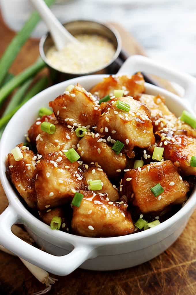 baked sesame chicken in a bowl with sesame seeds in a smaller bowl and green onions faded in the background.