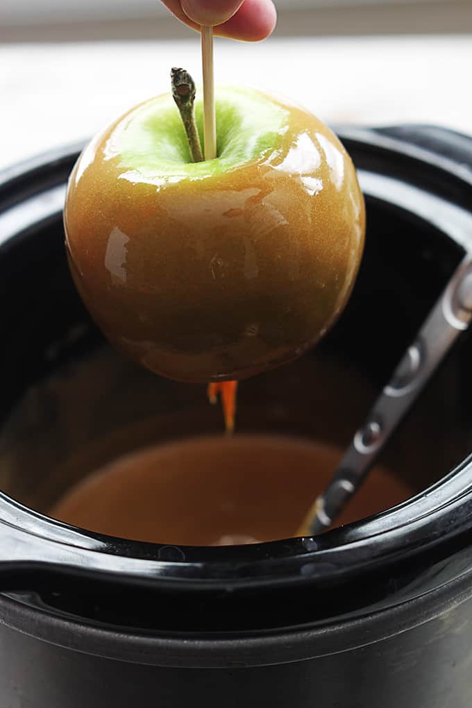 a caramel covered apple being held above a slow cooker filled with caramel fondue.