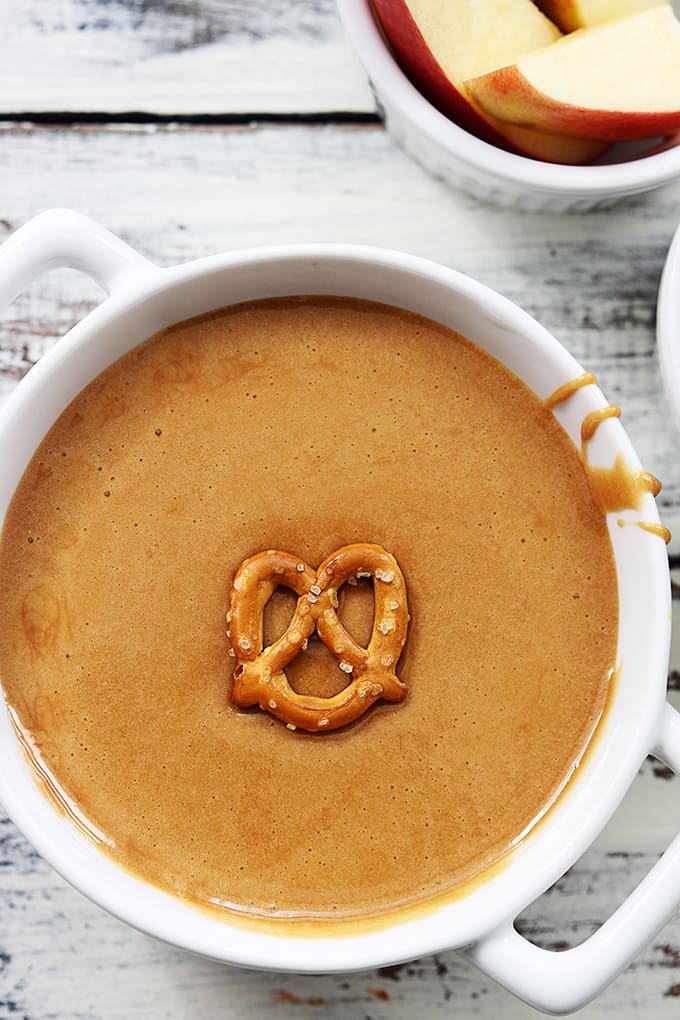 top view of a bowl of caramel fondue with a pretzel sitting on top and a bowl of apples on the side.