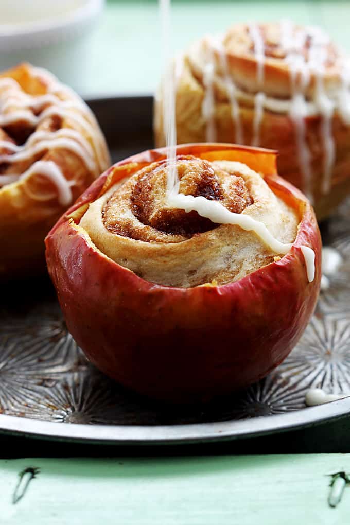 cinnamon roll stuffed baked apples on a serving tray with cream cheese frosting being poured on the front apple.