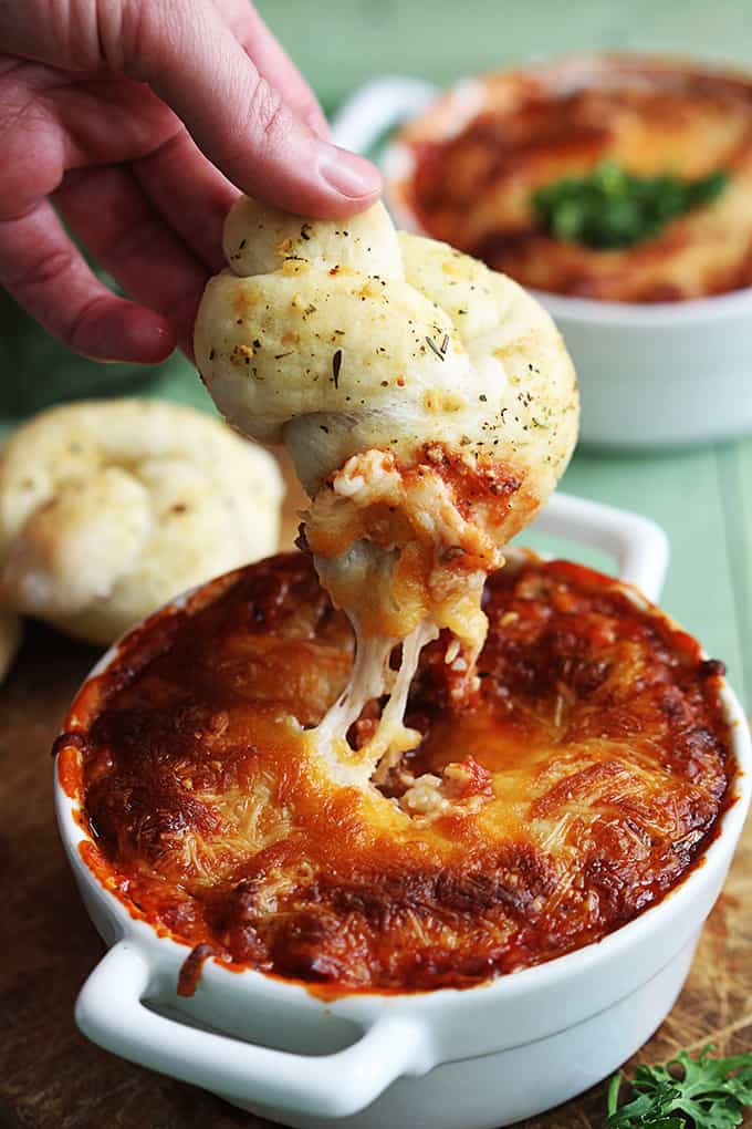 a hand holding a breaded knot just dipped in lasagna dip with the dip in a serving bowl underneath.