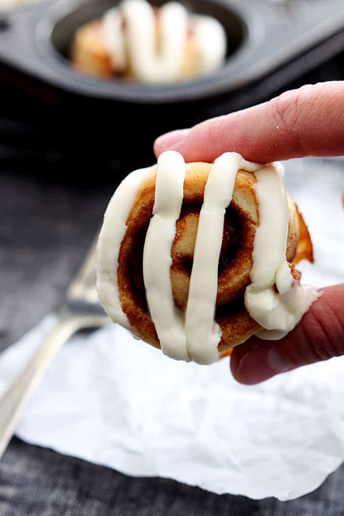 a hand holding a mini cinnamon roll topped with cream cheese frosting with a muffin tin of more rolls faded in the background.
