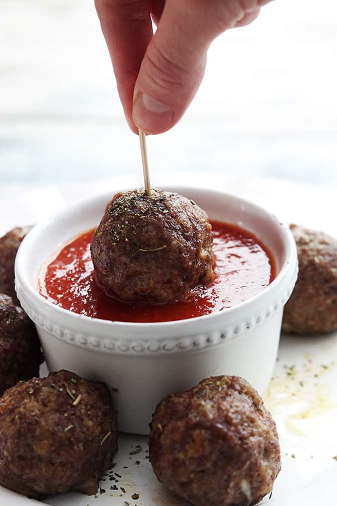 a hand holding a slow cooker mozzarella stuffed meatball on a toothpick dipping it in a bowl of marinara sauce with more meatballs around it.