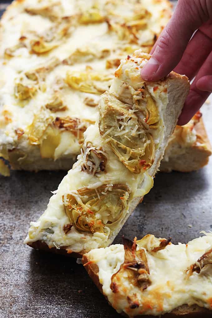 a hand picking up a slice of roasted garlic artichoke bread.