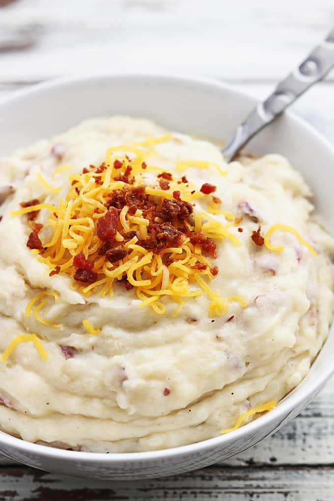 close up of slow cooker mashed potatoes with bacon bits and cheese on top with a spoon in a bowl.