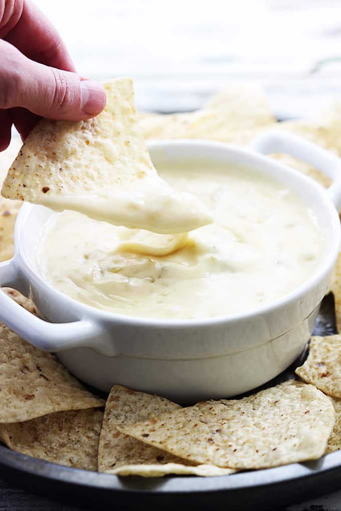 a hand holding a chip dipped in slow cooker queso blanco above a bowl of queso with more chips around it.