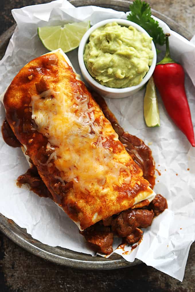 top view of a smothered Chile Colorado burrito on a round serving tray with a small bowl of guacamole, a red chili pepper, and slices of lime on the side.
