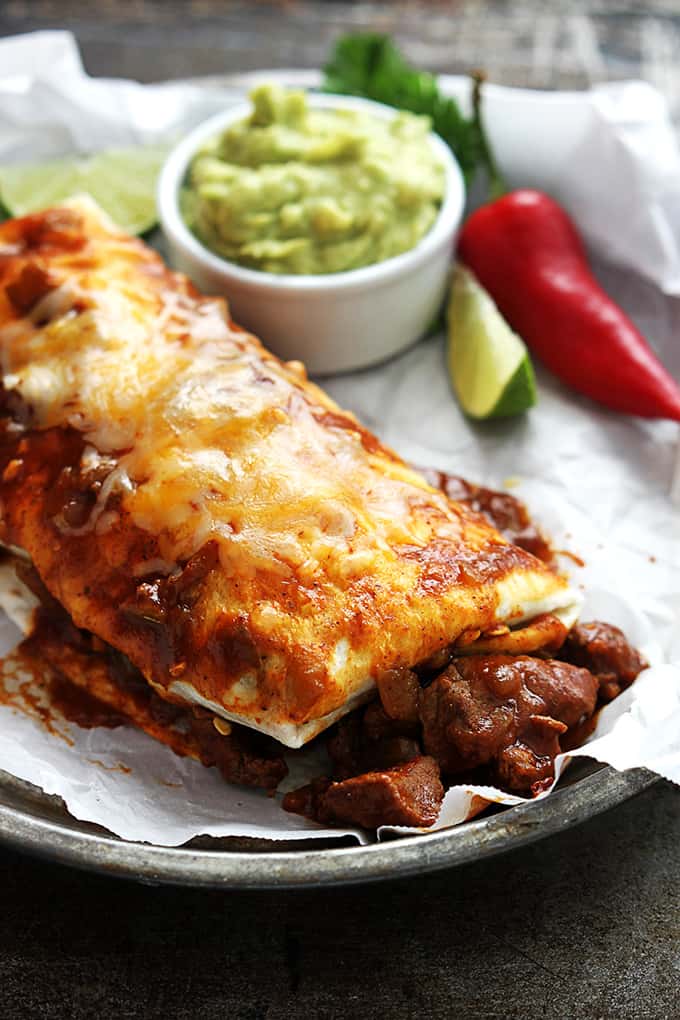 a smothered Chile Colorado burrito on a round serving tray with guacamole, a red chili pepper and a slice of a lime on the side.