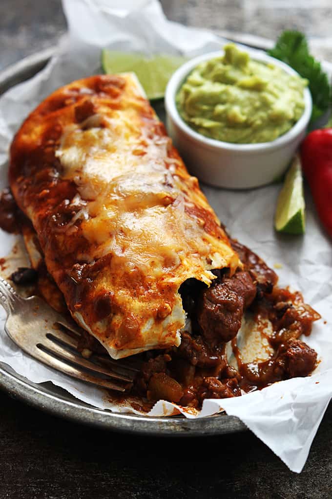 close up of a smothered Chile Colorado burrito with a fork, a bowl of guacamole, slices of lime and a red chili pepper on the side all in a round serving tray.