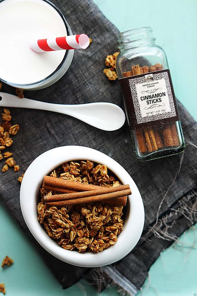 top view of cinnamon pumpkin granola in a bowl topped with cinnamon sticks with a jar of cinnamon sticks, a spoon and a glass of milk with a straw on the side.