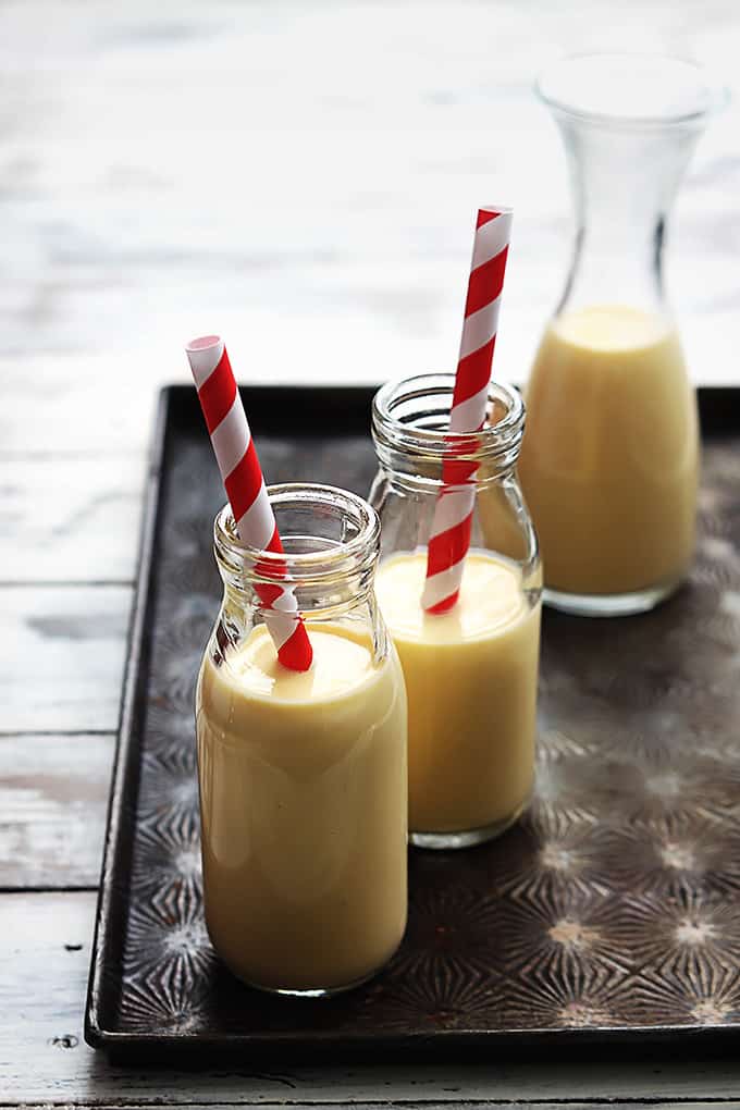 two glass jugs of milk filled with eggnog with two straws and a pitcher of more eggnog in the background.