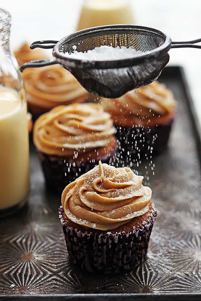 eggnog cupcakes topped with cinnamon cream cheese frosting with a sifter above sifting powdered sugar on top with glass milk gallons of eggnog on the side.