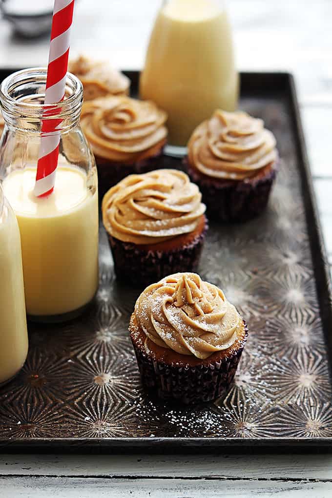 eggnog cupcakes topped with cinnamon cream cheese frosting and powdered sugar with glass milk jugs of eggnog on the side.
