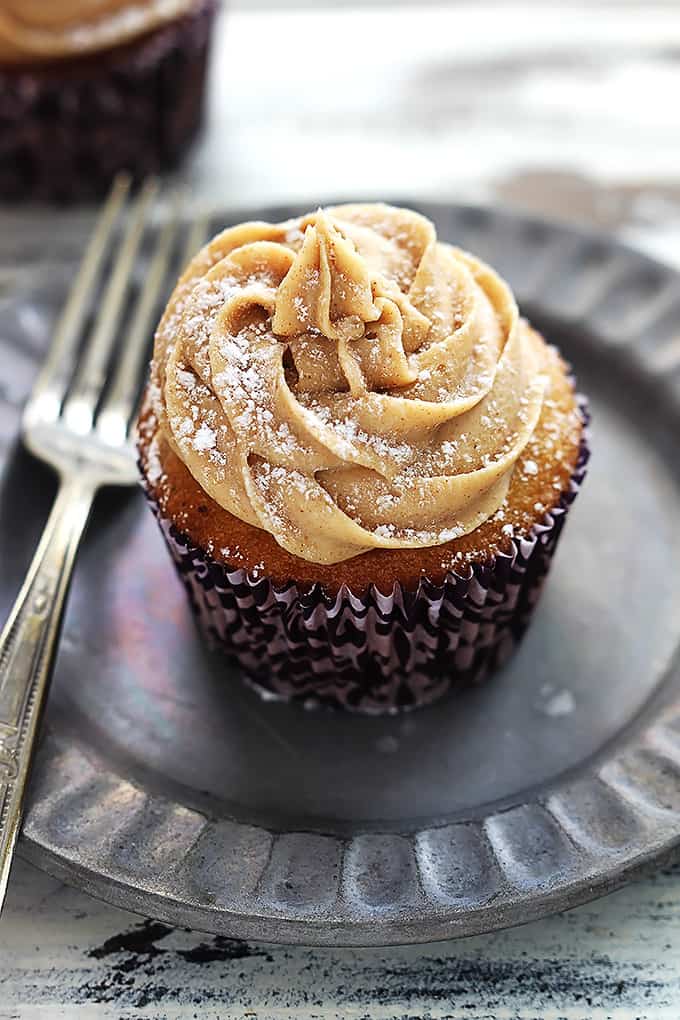an eggnog cupcake topped with cinnamon cream cheese frosting and powdered sugar with a fork on the side all on a plate.