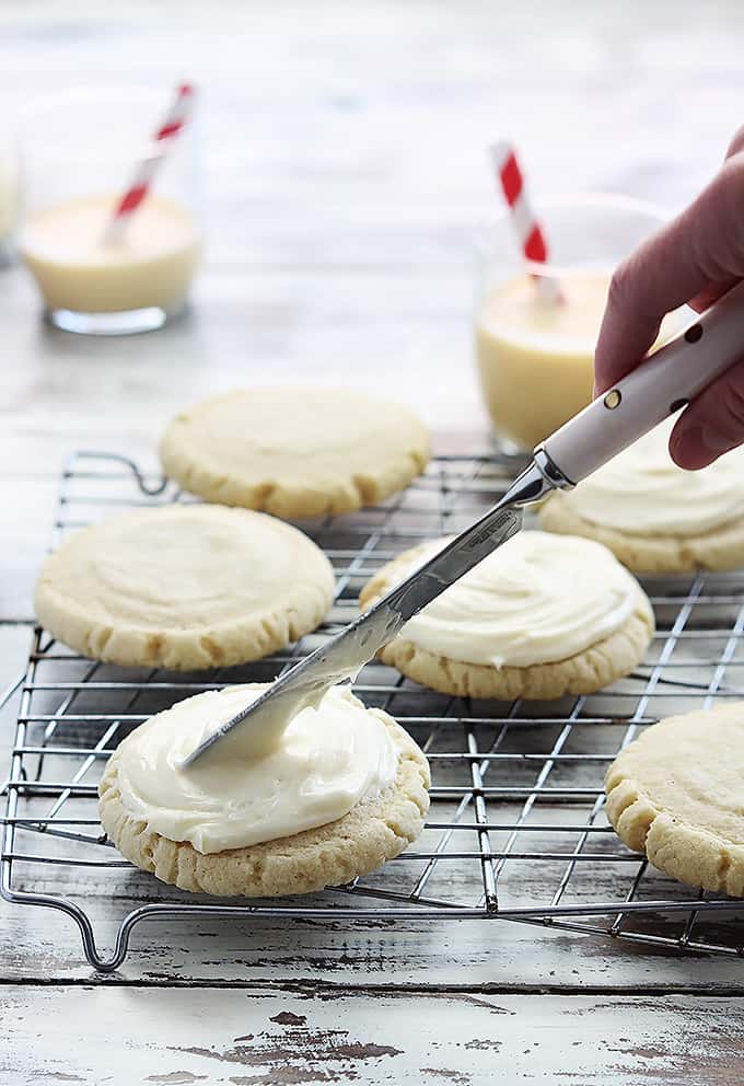 eggnog sugar cookies on a baking sheet with a hand holding a frosting knife frosting a cookie with glass of eggnog in the background.