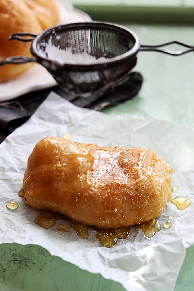 a fried scone topped with honey with a sifter above sifting powdered sugar on top.