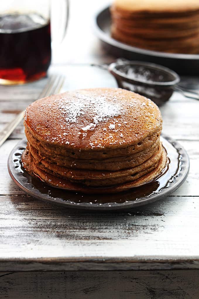 a stack of gingerbread pancakes on a plate with syrup and powdered sugar on top with a fork, sifter, syrup and more pancakes faded in the background.