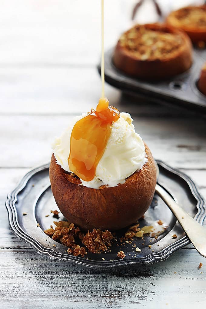a peach crisp stuffed baked peach on a plate topped with ice cream with caramel sauce being poured on top.