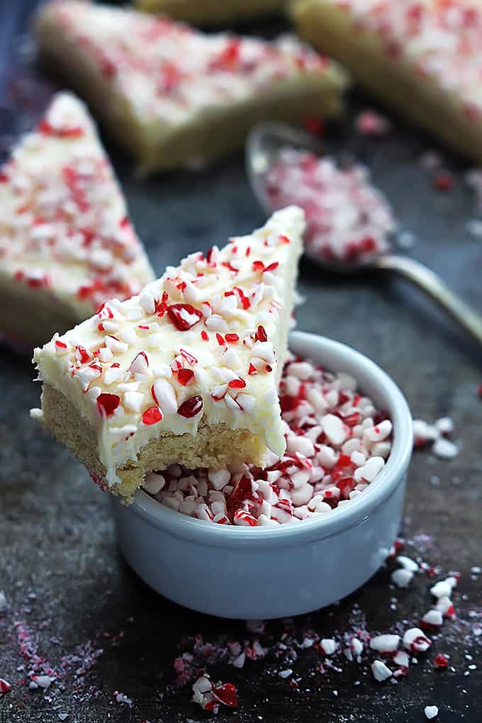 a peppermint crunch sugar cookie bar with a bite taken out of it sitting on top of a bowl of peppermint chunks with more bars and a spoon with peppermint chunks in the background.