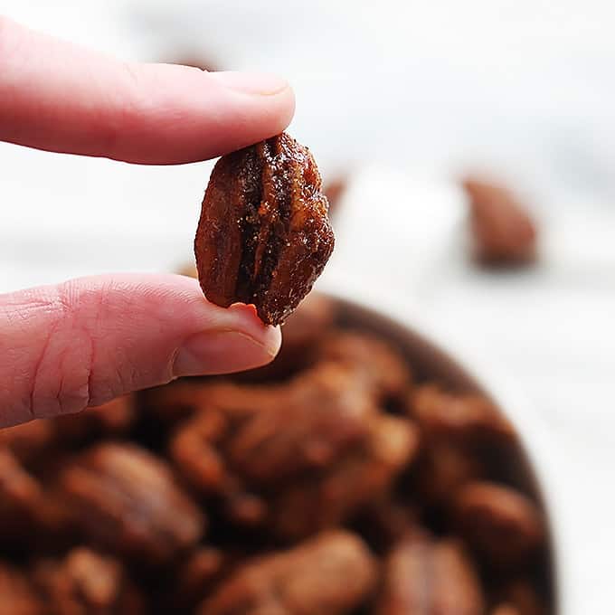 fingers holding up a slow cooker cinnamon pecans with more pecans faded in the background.
