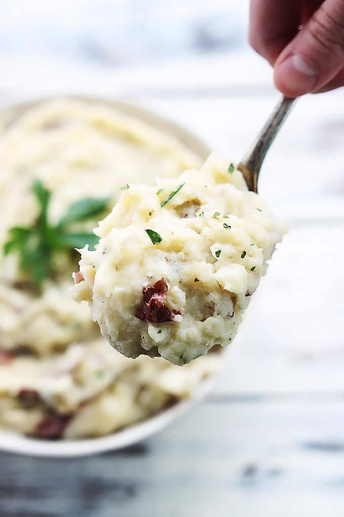 top view of a hand holding a scoop of slow cooker buttery garlic herb mashed potatoes on a spoon with a bowl of more potatoes faded in the background.