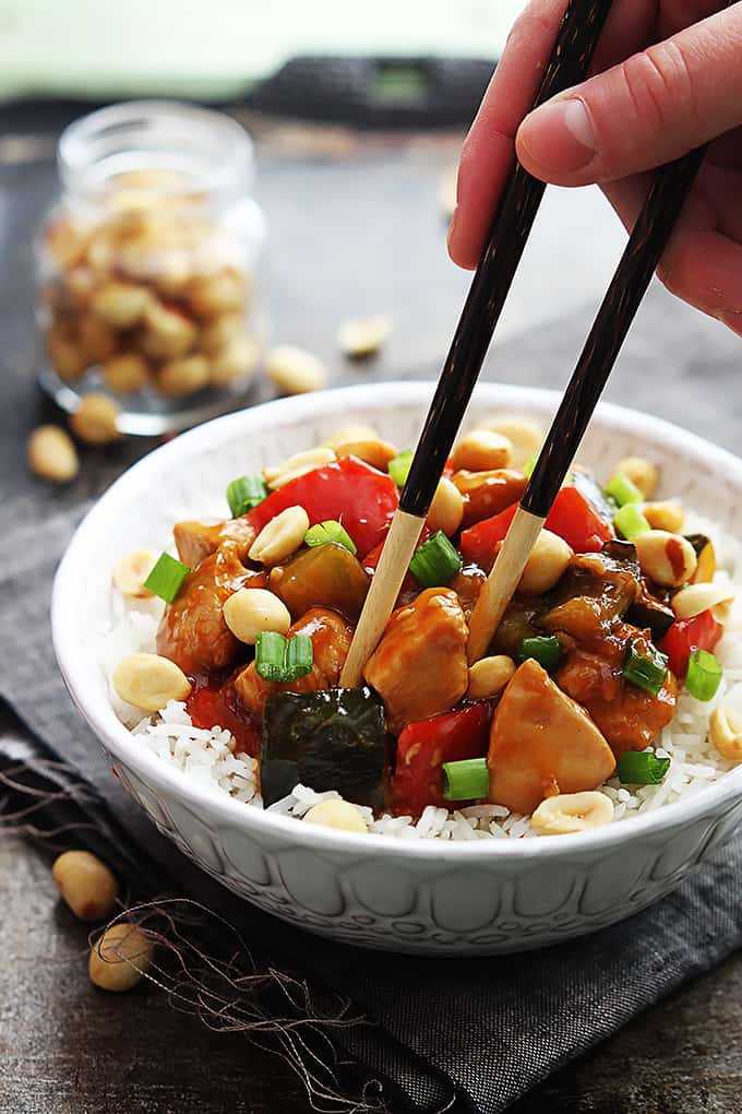 a hand holding chopsticks taking apiece of slow cooker kung pao chicken from a bowl of chicken on rice in a bowl topped with peanuts with a small jar of more peanuts on the side.