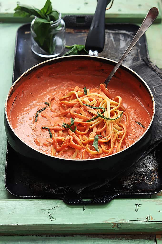 creamy tomato basil fettuccini with a swerving spoon in a skillet on a vintage baking sheet with a jar of garnish in the background.