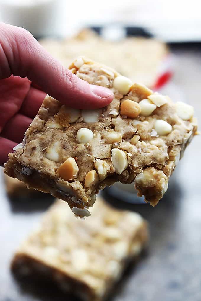 a hand holding a white chocolate chip macadamia nut blondie with a bite taken out with more blondies faded in the background.