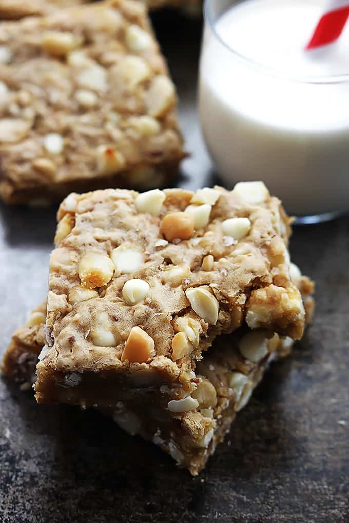 stacked white chocolate chip macadamia nut blondies with a bite missing out of the top one with more blondies and a glass of milk in the background.