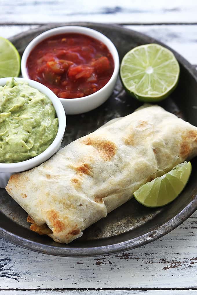 a baked chicken chimichanga on a round serving tray with limes and guacamole and salsa in small bowls on the side.
