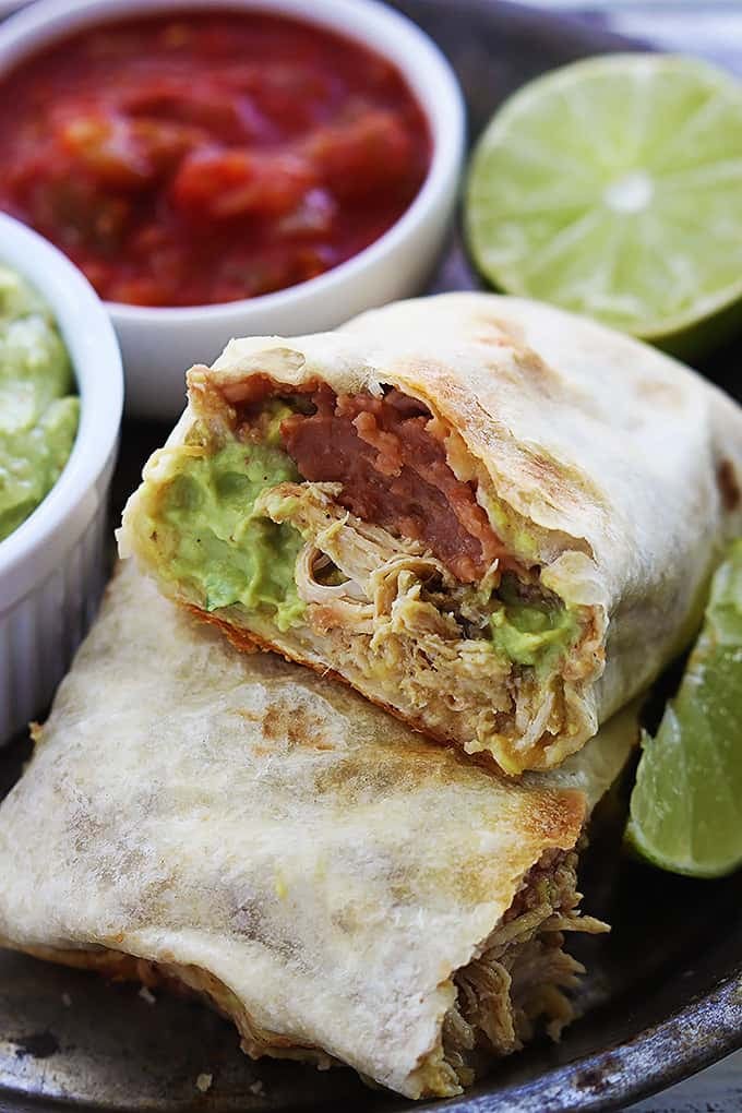 close up of a baked chicken chimichanga cut in half with guacamole, limes, and salsa on the side.
