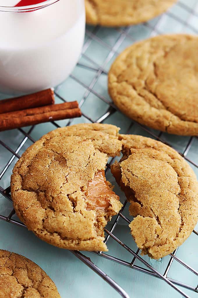 close up of a caramel stuffed snickerdoodle cookie broken in half with more cookies, cinnamon sticks and a glass of milk on the side all on a cooling rack.