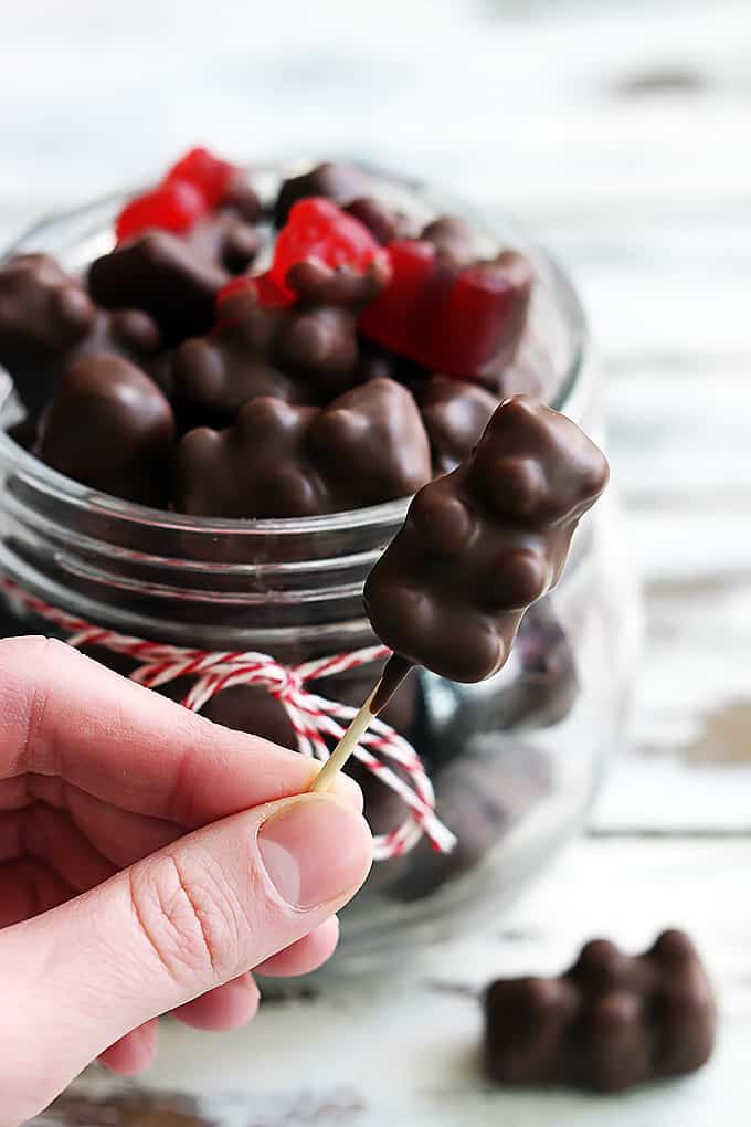 a hand holding a chocolate covered cinnamon bear on a toothpick with a jar of more bears in the background.