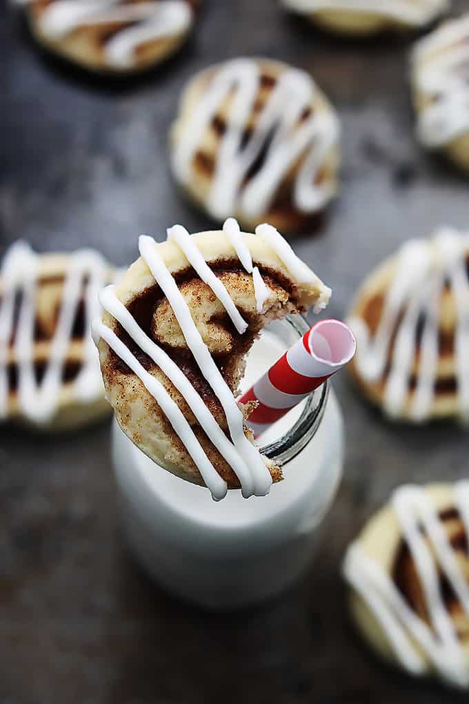 a cinnamon roll cookie with a bite taken out of it sitting on top of a glass jar of milk with more cookies on the side.