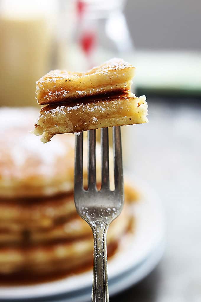 a bite of eggnog pancakes on a fork with a stack of eggnog pancakes on a plate and a glass bottle of eggnog faded in the background.