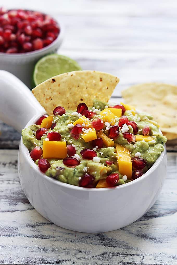 a bowl of mango pomegranate guacamole with a chip dipped in it with more chips, half of a lime, and a bowl of pomegranate in the background.