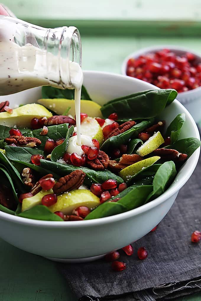 pomegranate pear & pecan salad in a bowl with poppyseed dressing being poured on top with a bowl of pomegranates in the background.