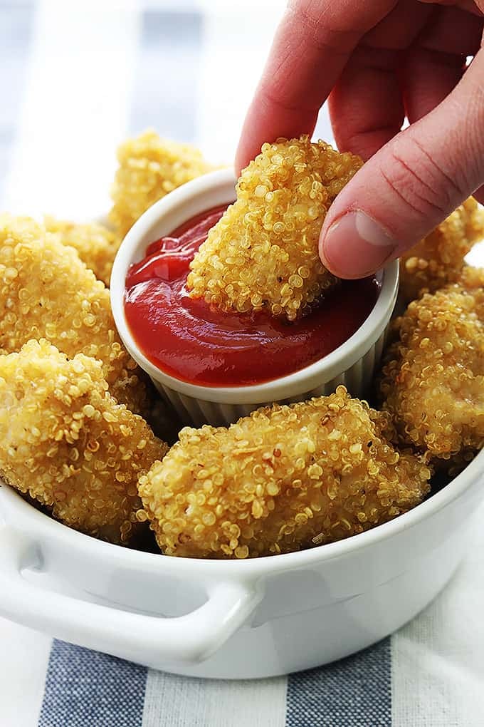 a hand dipping a quinoa chicken nugget in a small dipping bowl of ketchup in the middle of a bowl full of more nuggets.