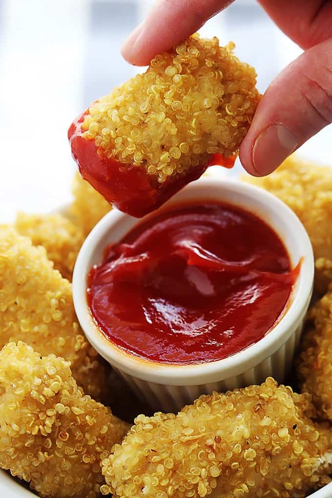 a hand holding a quinoa chicken nugget dipped in ketchup above a bowl of more nuggets with a small dipping bowl of ketchup in the middle.