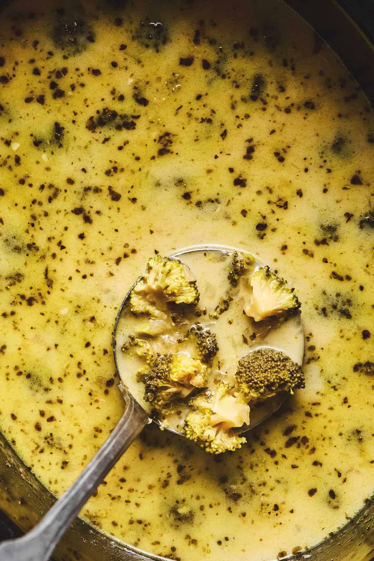 Close up overhead of a label lifting up some slow cooker broccoli cheddar cheese soup from a slow cooker full of soup.