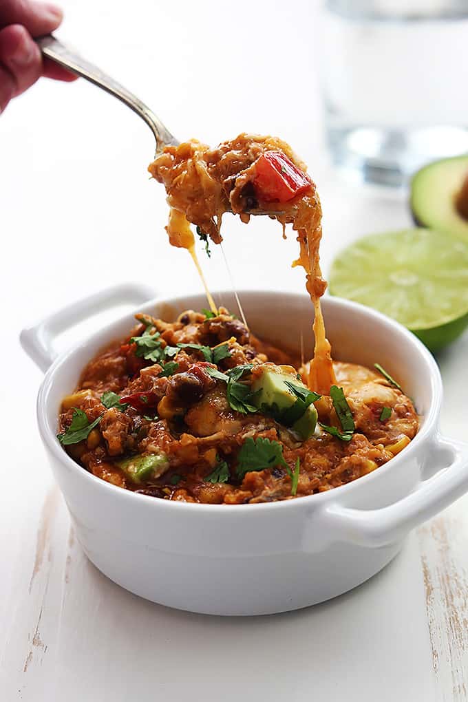 a hand holding a scoop of slow cooker enchilada quinoa on s spoon above a bowl of more quinoa with half of a lime and avocado in the background.