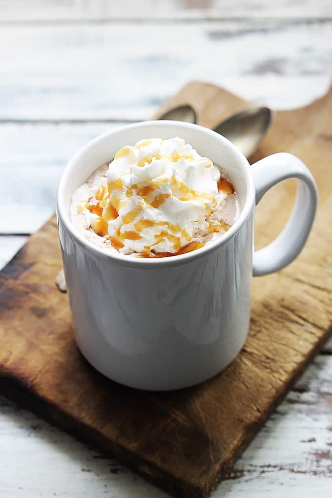 slow cooker salted caramel hot chocolate in a mug topped with whipped cream and caramel on a cutting board.