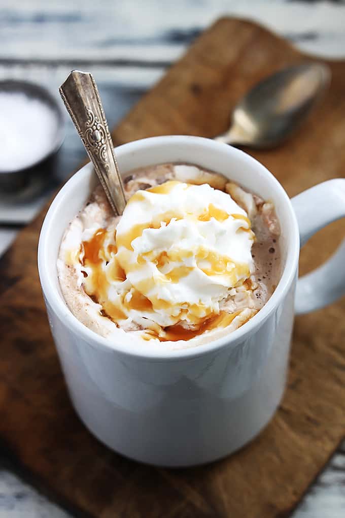 slow cooker salted caramel hot chocolate in a mug with a spoon topped with whipped cream and caramel on a wooden cutting board with another spoon and a bowl of salt faded in the background.