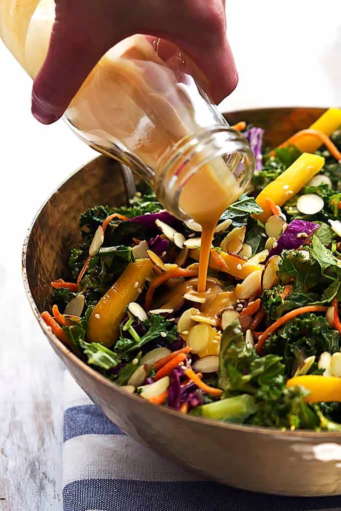 a hand pouring creamy peanut dressing on top of Asian kale salad in a salad bowl.