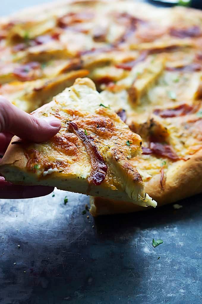 a hand picking holding a slice of bacon chicken ranch pizza with a bite missing in front of the rest of the pizza.